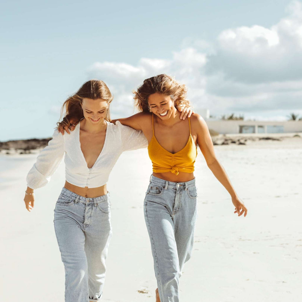 two women walk on the beach, smiling, wearing light wash denim